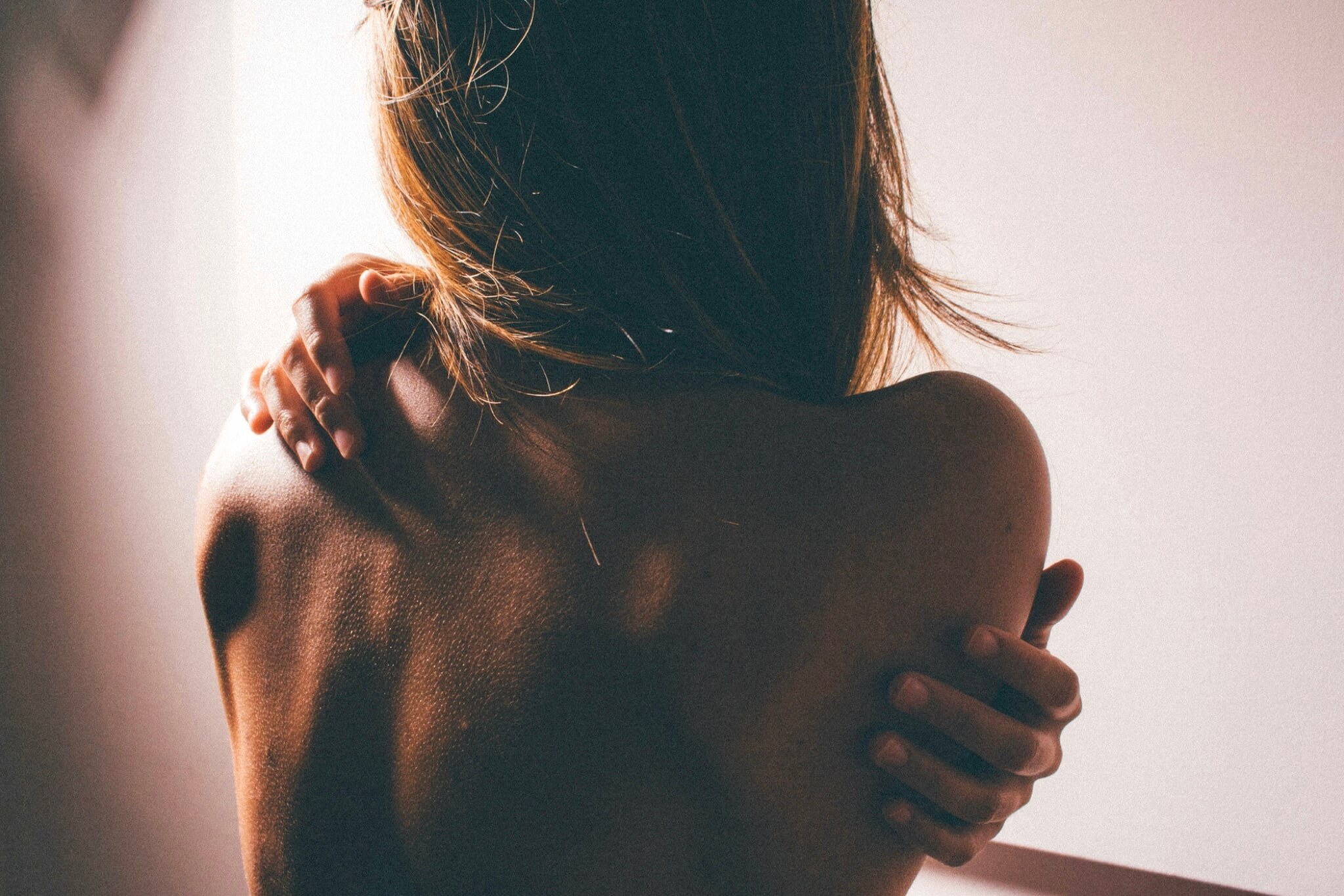 A figure turned away, holding her own shoulder in soft warm light
