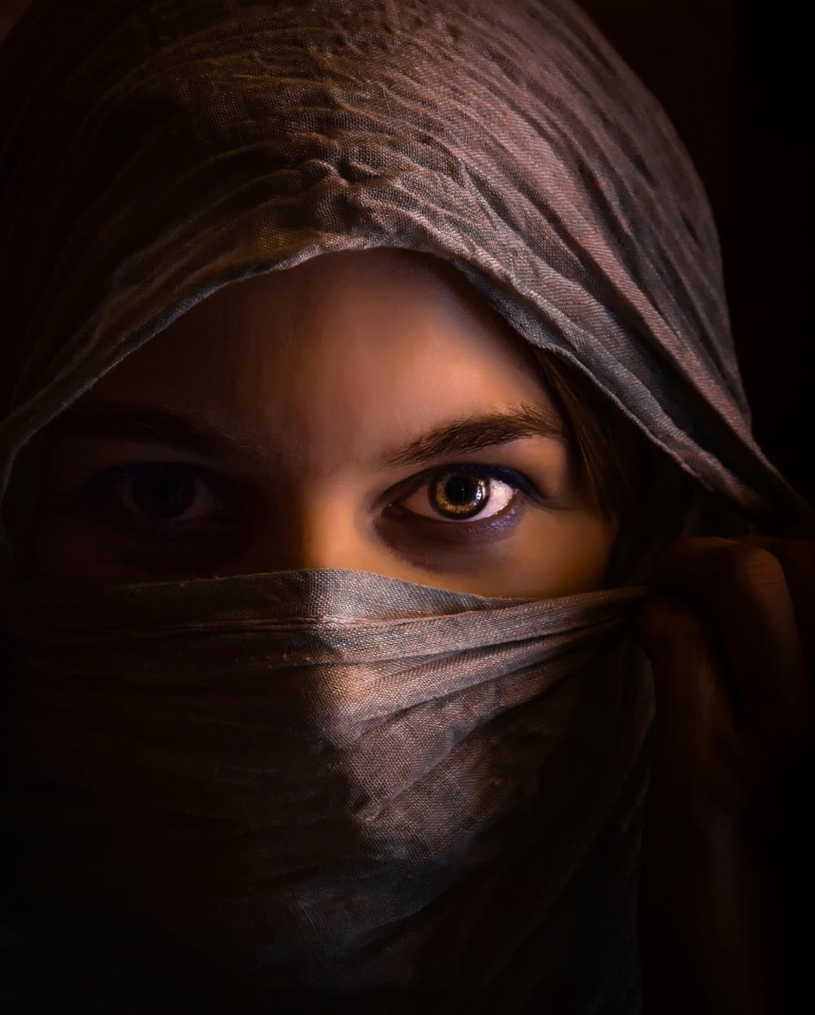 Veiled face with luminous, watchful eyes emerging from shadow