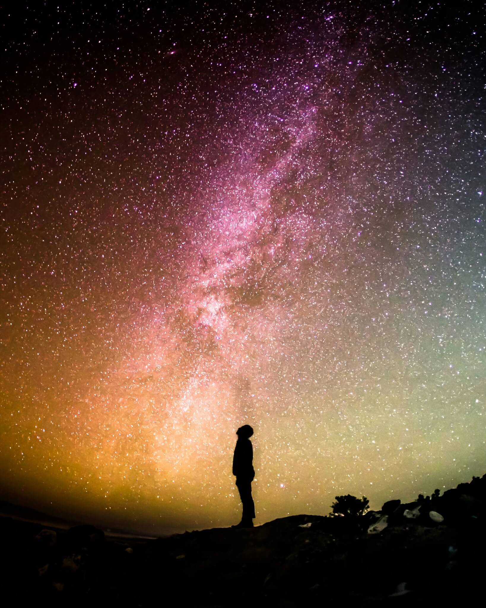 A lone figure standing beneath a vast, luminous, colour-drenched Milky Way